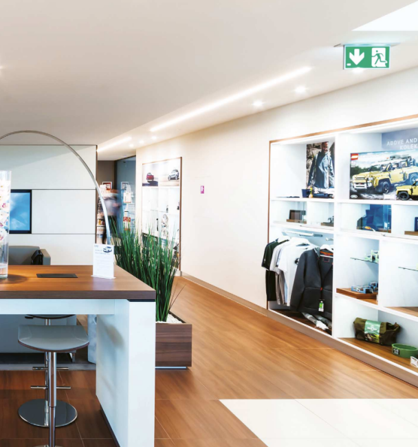 Čistý moderní interiér showroomu s vestavěnými vitrínami a nábytkem z tmavého dřeva, osvětlený LED lištami.