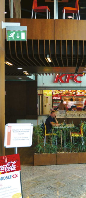 Bezpečnostní značení nad únikovým východem ve foodcourtu s viditelným logem KFC a zákaznickou zónou.